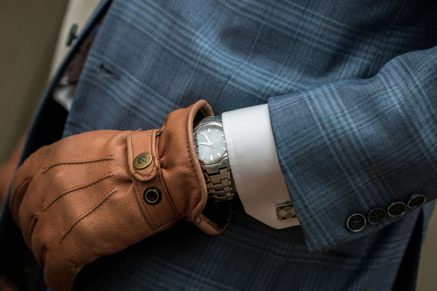 Close-up of a stylish man in a blue suit and brown gloves checking the time on a silver wristwatch.